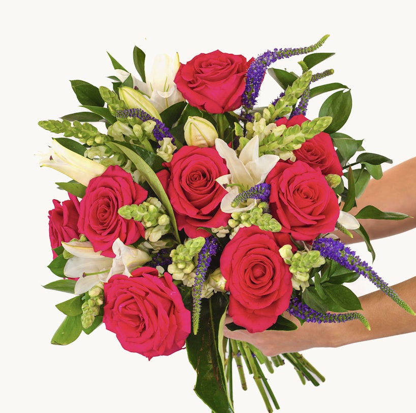 Vibrant bouquet of flowers featuring red roses, white lilies, and pink blooms with greenery and hints of purple flowers, held against a white background.