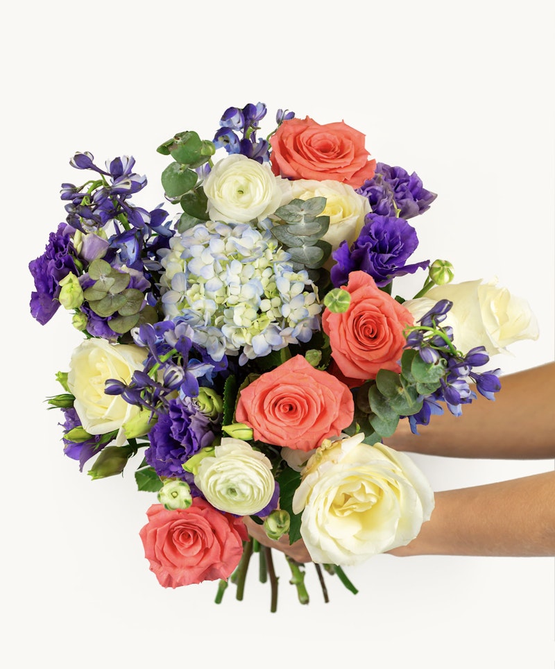 Hands presenting a vibrant bouquet of flowers with a mix of pink, white and purple blooms and lush greenery on a white background, perfect for celebrations or decor.