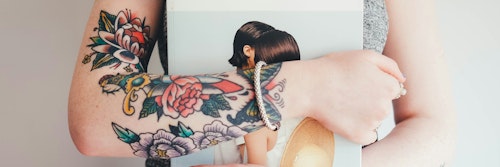 Close-up of an arm with vibrant traditional tattoos, including a red rose and a bird, with a person holding a book in the background.