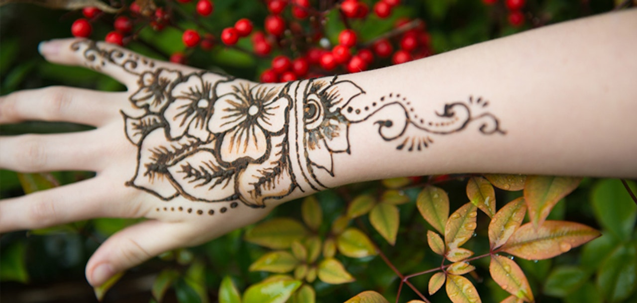 A hand adorned with intricate henna tattoo designs featuring floral patterns rests against a background of green leaves and vibrant red berries.
