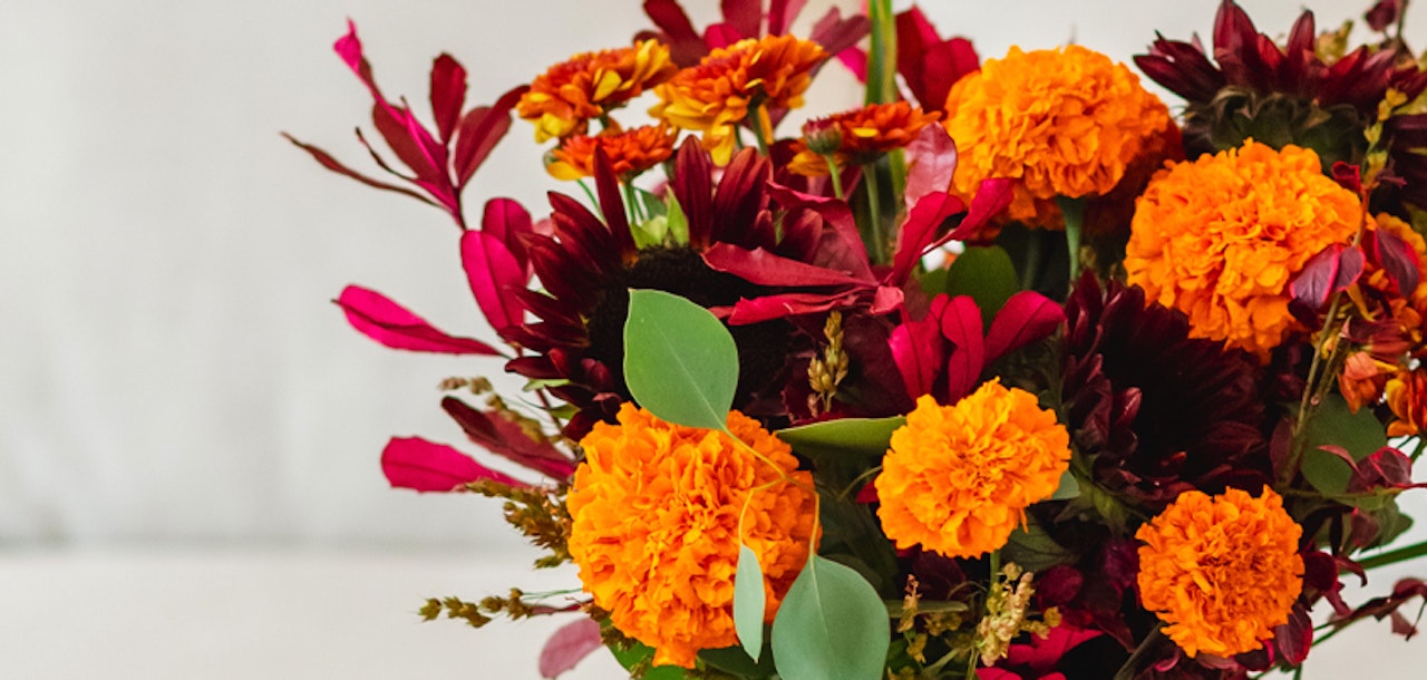 Vibrant bouquet of autumn flowers featuring orange marigolds and deep red blossoms with green foliage on a white background, symbolizing fall's vivid colors.