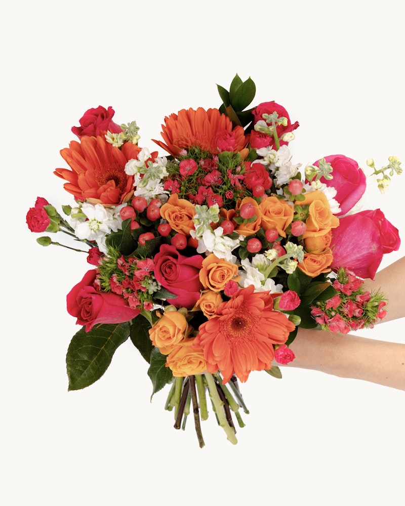 A person holding a vibrant bouquet of flowers featuring orange gerberas, pink roses, and various greenery against a white background, perfect for special occasions.