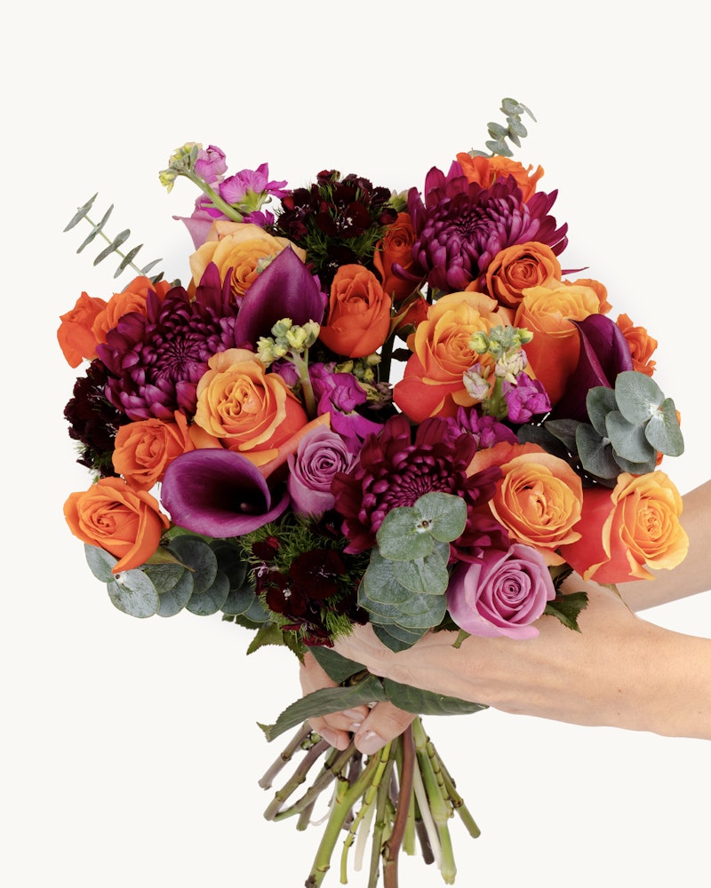 A vibrant bouquet of flowers, including orange roses, purple calla lilies, and deep red blooms, held against a light background, showcasing a variety of colors and textures.
