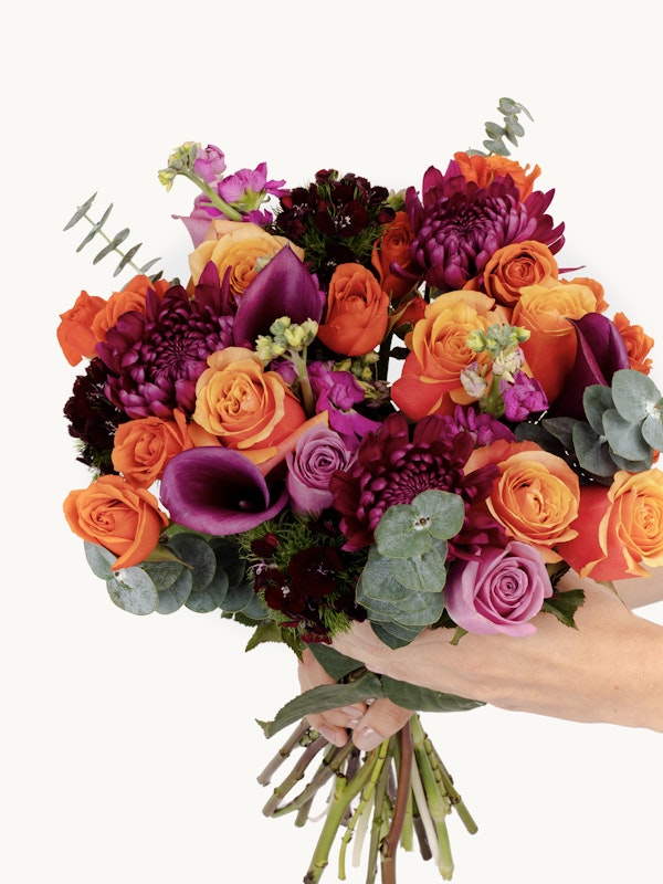 A vibrant bouquet of flowers, including orange roses, purple calla lilies, and deep red blooms, held against a light background, showcasing a variety of colors and textures.