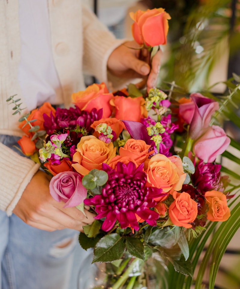 Person in a cream cardigan arranging a vibrant bouquet of flowers, including orange roses, pink calla lilies, and purple blooms, with green foliage accents.