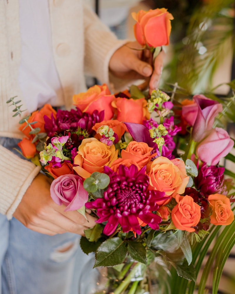 Person in a cream cardigan arranging a vibrant bouquet of flowers, including orange roses, pink calla lilies, and purple blooms, with green foliage accents.