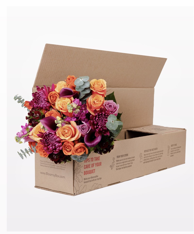 Vibrant bouquet of orange roses, purple flowers, and green leaves packaged neatly in an open cardboard box from BloomsyBox.com, with care instructions on the side.