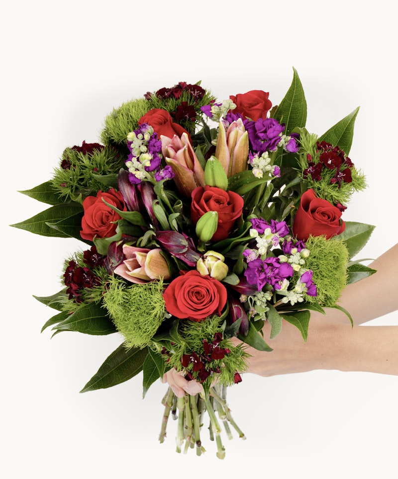 A person's hand holding a vibrant bouquet of flowers, featuring red roses, purple lilies and accents, with lush greenery on a white background.