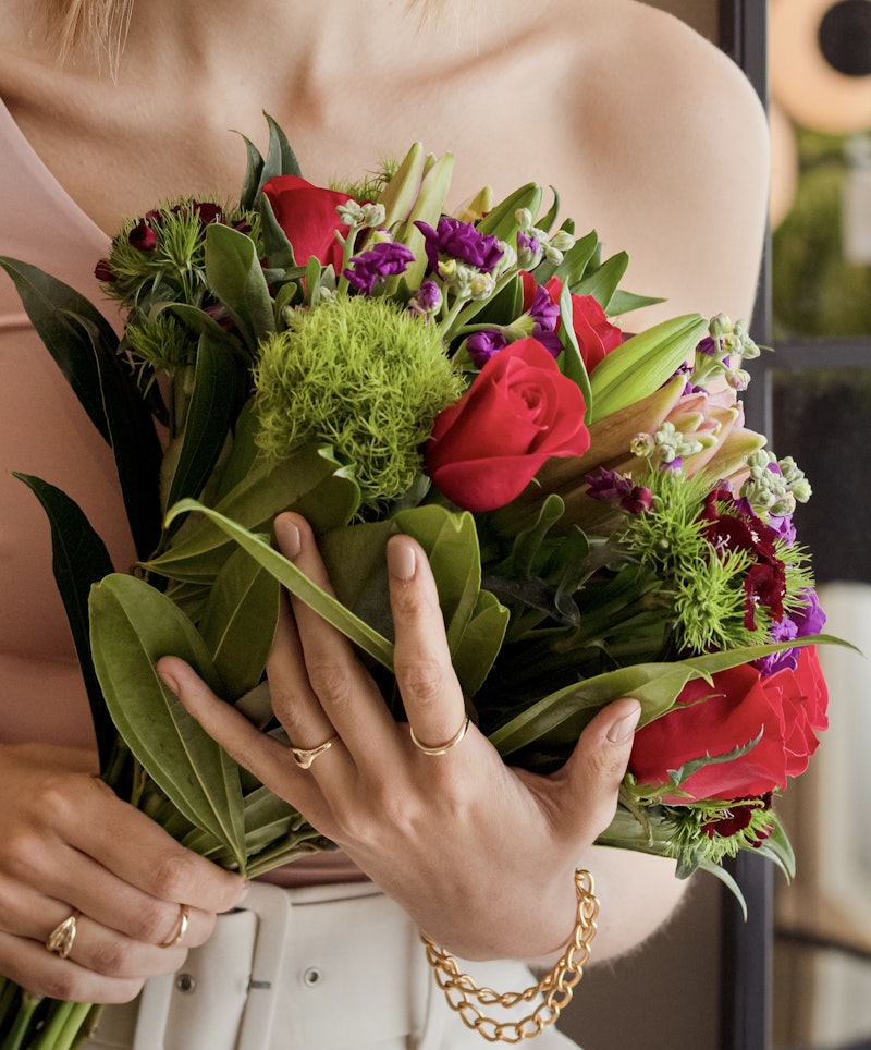 A person wearing a gold chain bracelet and a ring holding a vibrant bouquet with red roses, purple flowers, and green foliage, suggesting a special occasion or a gift gesture.