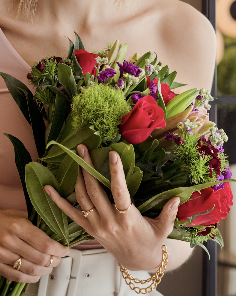 A person wearing a gold chain bracelet and a ring holding a vibrant bouquet with red roses, purple flowers, and green foliage, suggesting a special occasion or a gift gesture.