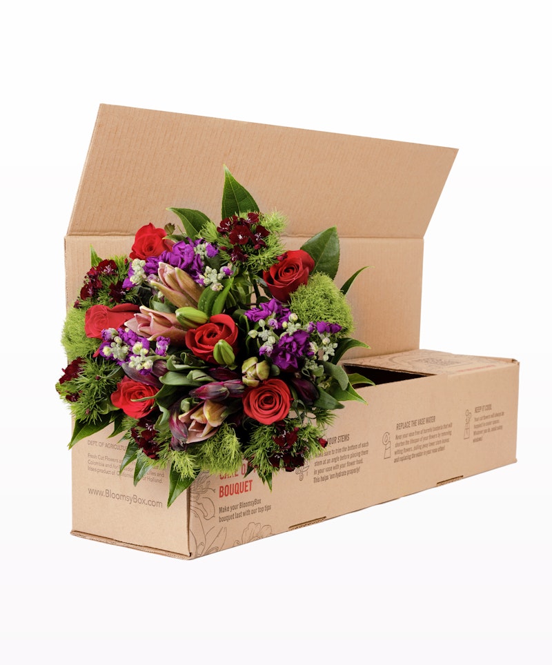 Vibrant bouquet of red roses, purple flowers, and greenery emerging from a cardboard delivery box with the label "BloomyBox.com BOUQUET" on a white background.