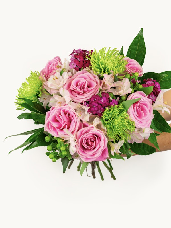 A vibrant bouquet of pink roses, green chrysanthemums, and various purple flowers held by a person against a white background, showcasing a mix of blossoms and foliage.