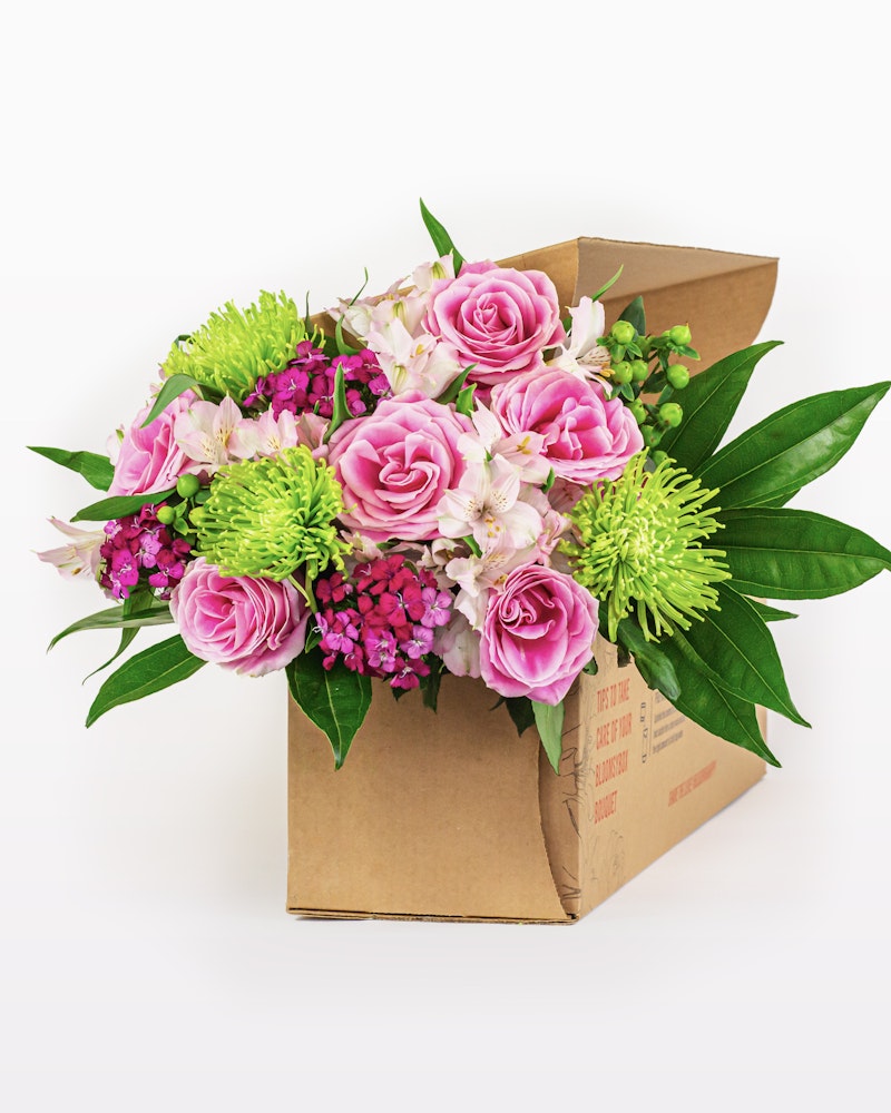 Vibrant bouquet of fresh flowers with pink roses, green chrysanthemums, and purple accents arranged in a cardboard delivery box against a white background.