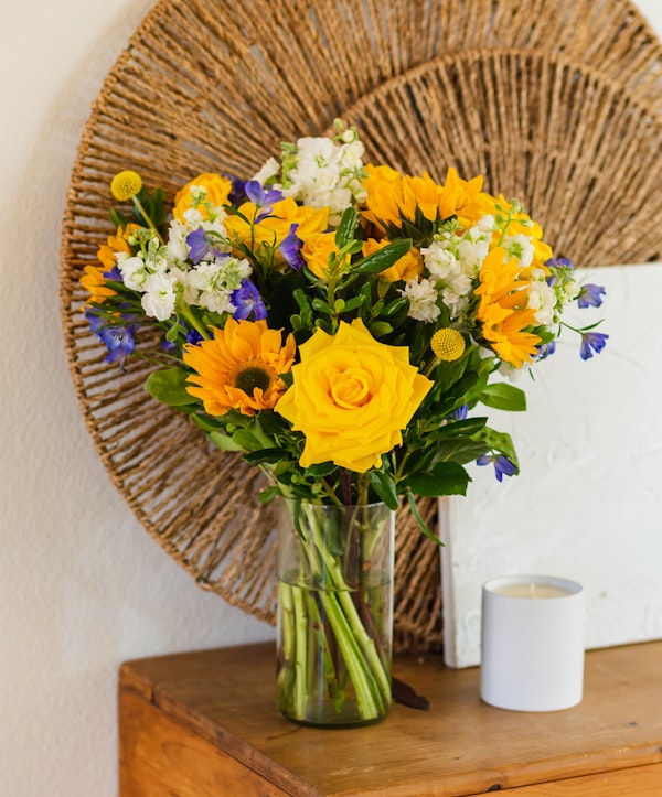 Bright bouquet of yellow roses and sunflowers with purple and white accents in a clear vase on a wooden table against a textured wall with a round wicker decoration.