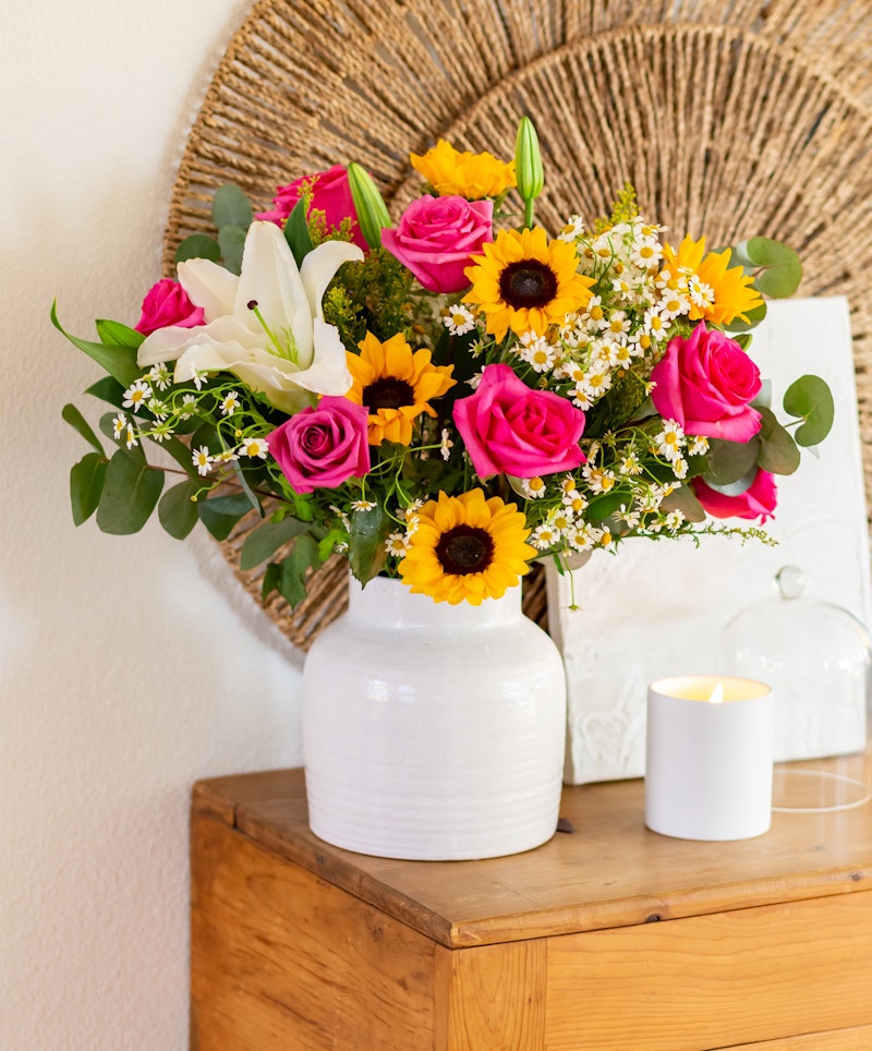 Bright bouquet of sunflowers, pink roses, and white lilies in a white vase on a wooden table, with a wicker decoration and a lit candle nearby, evoking a warm, cozy ambiance.