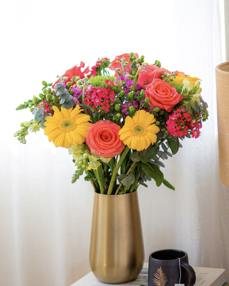 Vibrant bouquet of flowers featuring sunflowers, pink roses, and mixed greenery in a golden vase on a table beside a coffee cup with a fern design.