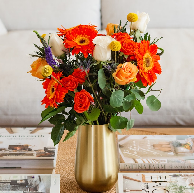 A vibrant bouquet of orange gerberas, white tulips, and roses in a gold vase, set on a coffee table amidst various home decor magazines.
