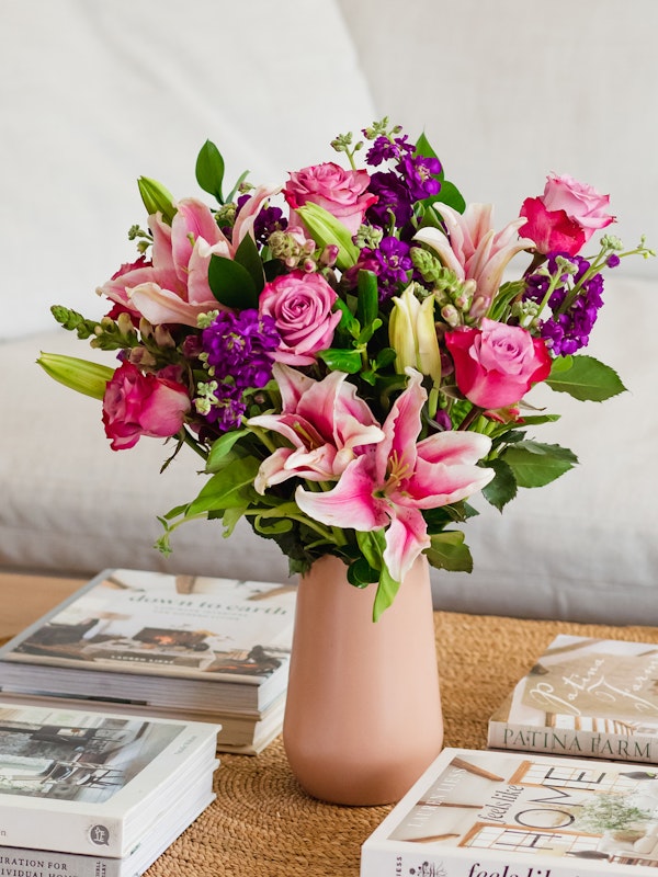A vibrant bouquet of pink roses, lilies, and purple flowers in a blush vase, placed on a stack of hardcover books with a cozy beige cushion background.