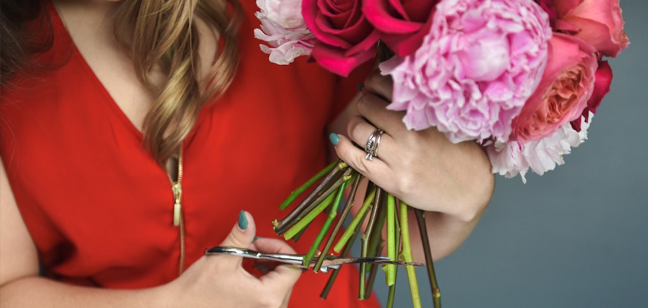 Woman in a red dress trims the stems of a bouquet of vibrant pink peonies with scissors, focusing on flower arrangement and gardening.