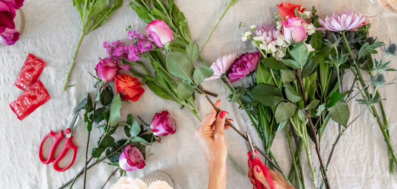 A flat lay of vibrant assorted flowers being arranged on a white cloth, with a person's hand trimming stems and floral scissors on display, crafting a bouquet.