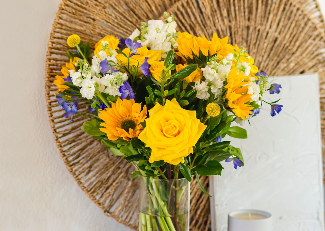 Bright and cheerful bouquet featuring yellow roses, sunflowers, and blue blossoms in a glass vase.