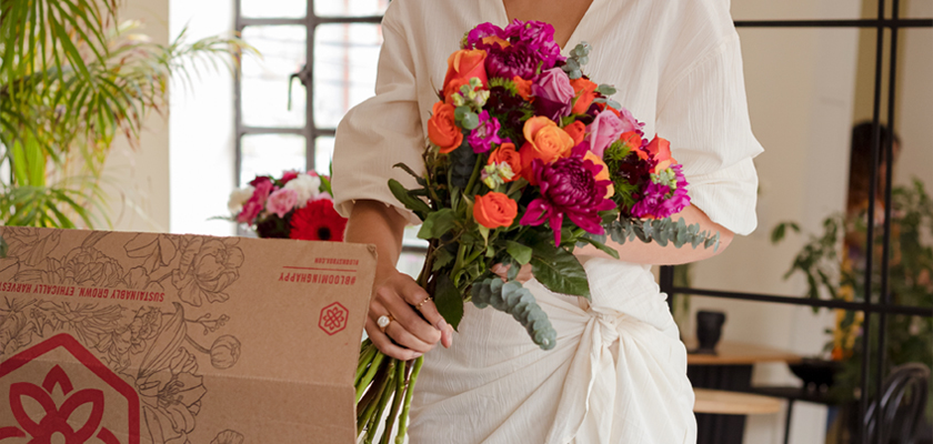 A woman in a white outfit holds a vibrant floral bouquet with roses and peonies.