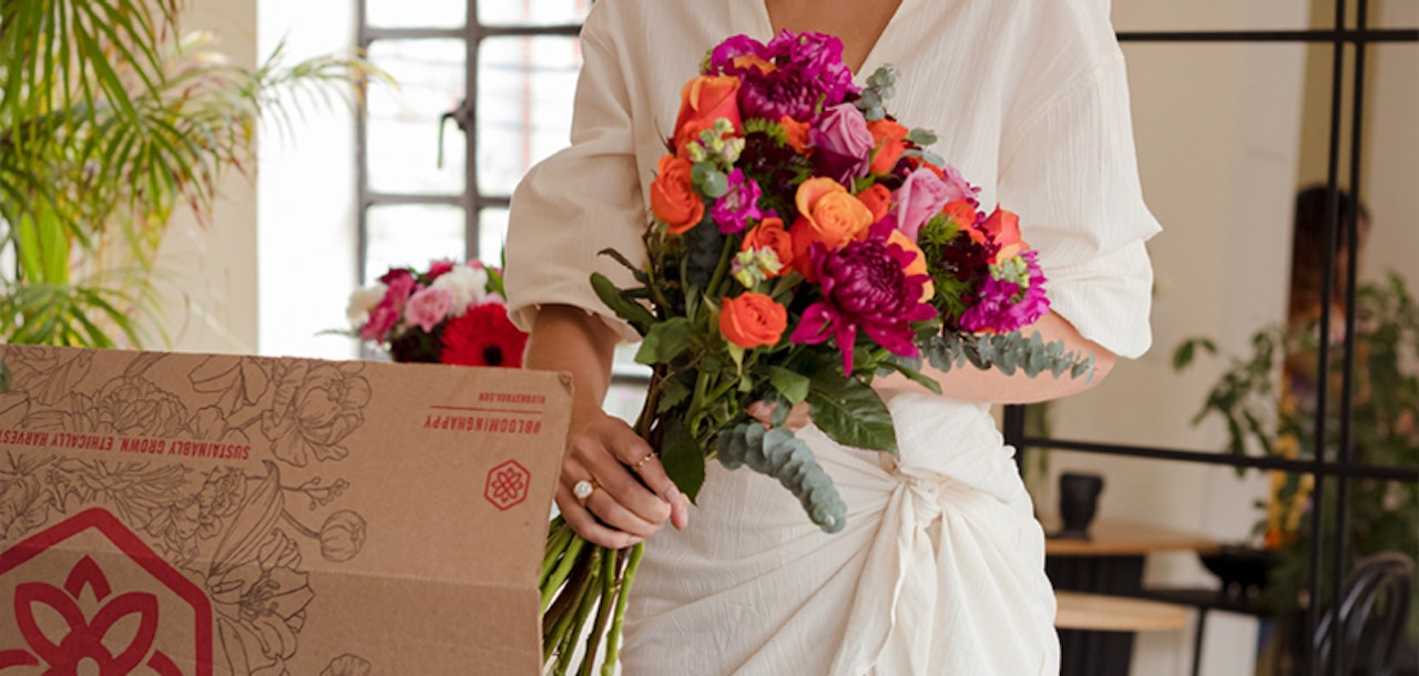 Woman in a white blouse holding a vibrant bouquet of flowers, standing next to a cardboard box on a table in a room with natural light.