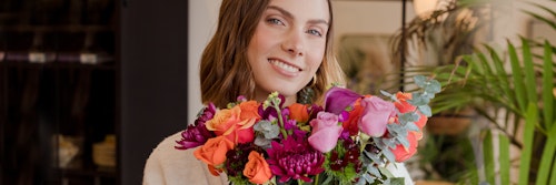 Smiling woman holding a vibrant bouquet of flowers inside a cozy room with green plants and a blurred background, conveying a sense of warmth and happiness.
