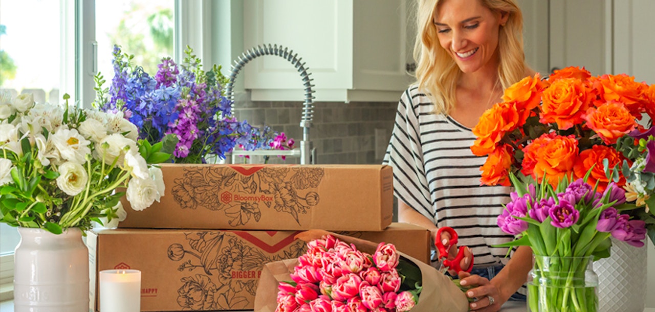 Smiling woman arranging a vibrant bouquet of pink roses in her kitchen, with boxes of flowers and assorted colorful blooms on the countertop.