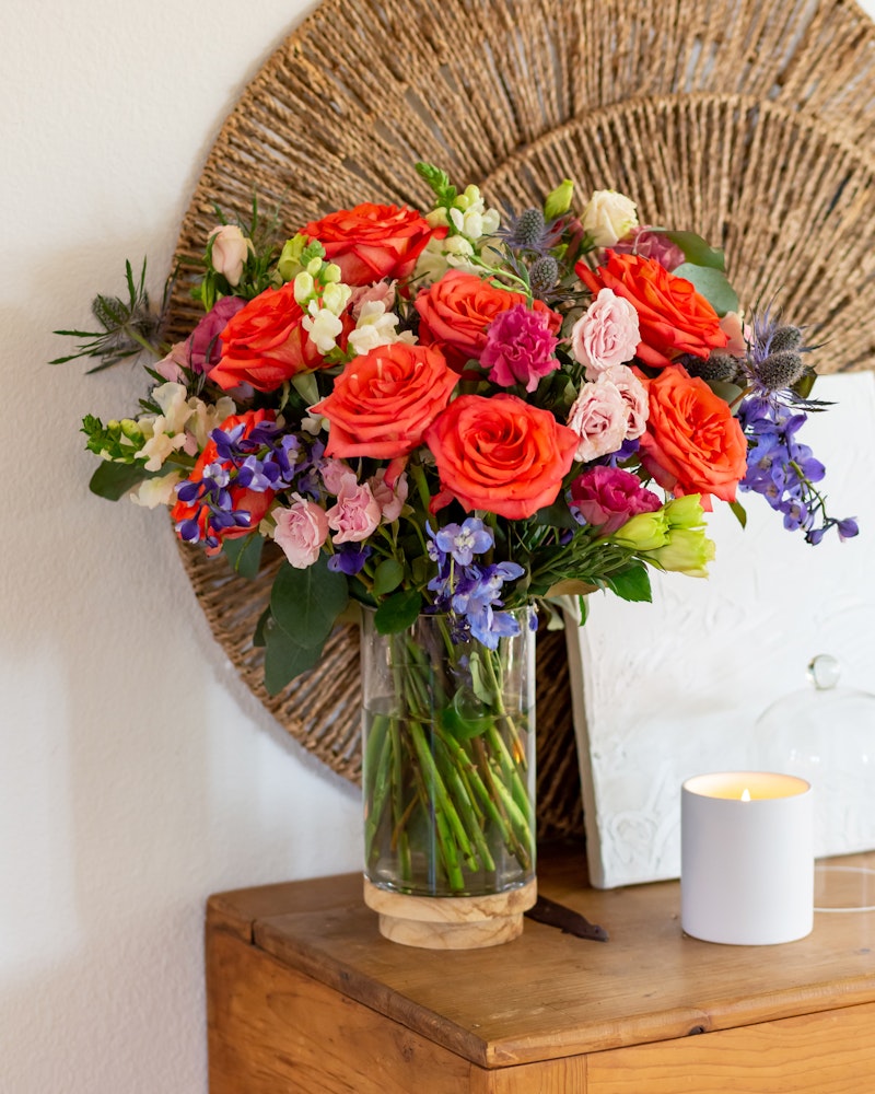 Vibrant bouquet of red roses and purple flowers in a clear vase on a wooden table beside a lit candle, with a white frame and wicker decoration in the background.