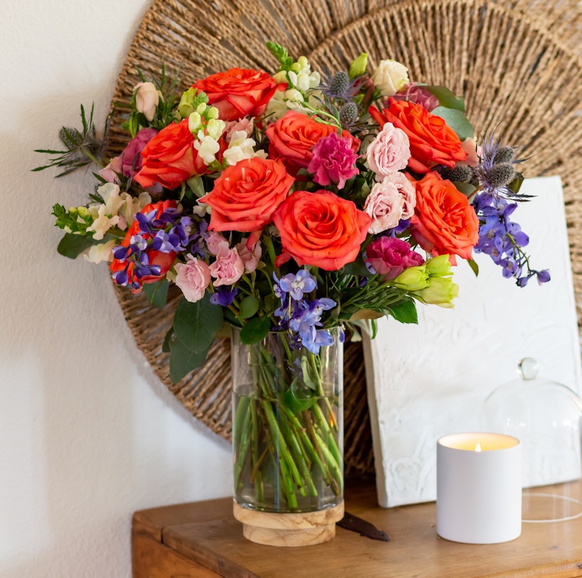 Vibrant bouquet of red roses and purple flowers in a clear vase on a wooden table beside a lit candle, with a white frame and wicker decoration in the background.