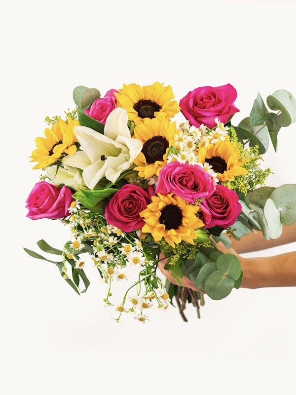 Two hands holding a vibrant bouquet of flowers including sunflowers, pink roses, white lilies, and small daisies against a white background.