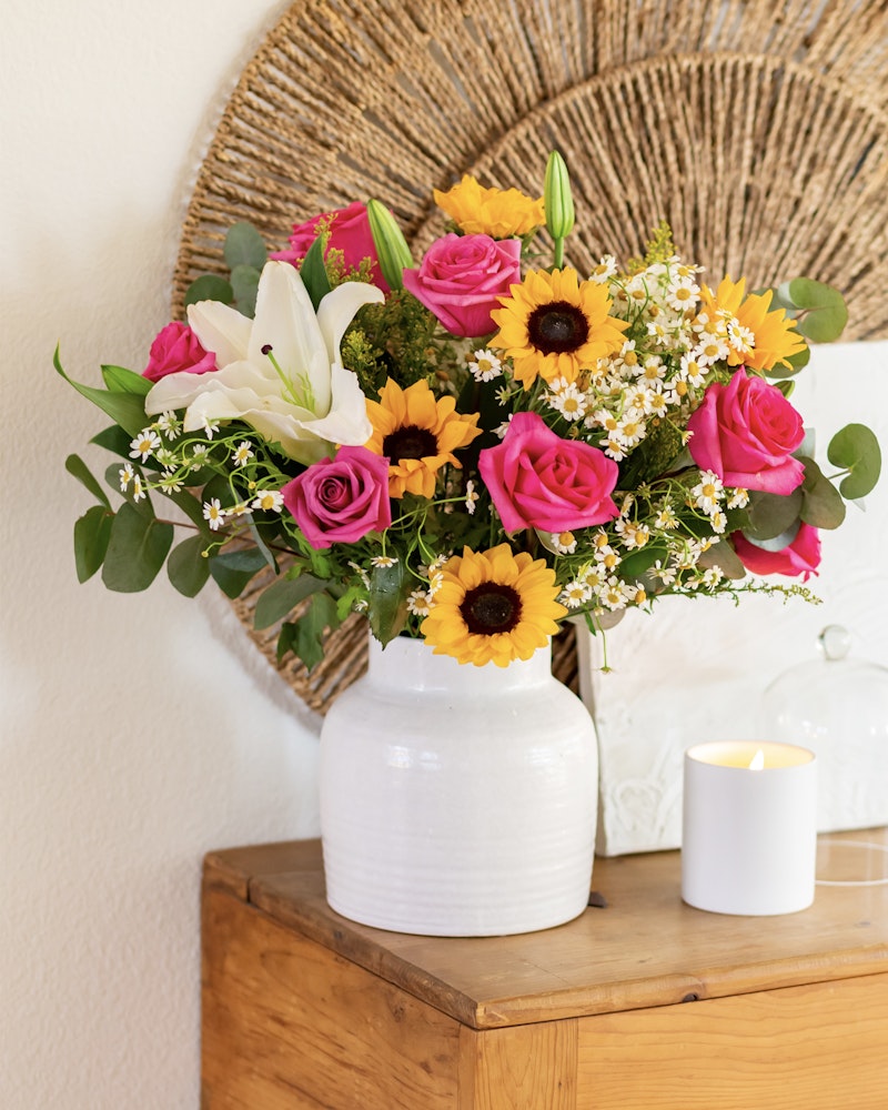 Vibrant floral arrangement in a white vase featuring sunflowers, pink roses, and lilies on a wooden side table, with a lit candle and woven wall decor in the background.