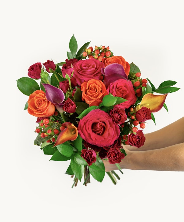 A lush bouquet with a mix of vibrant red roses, orange roses, and purple calla lilies, accented with green foliage, presented against a white background.