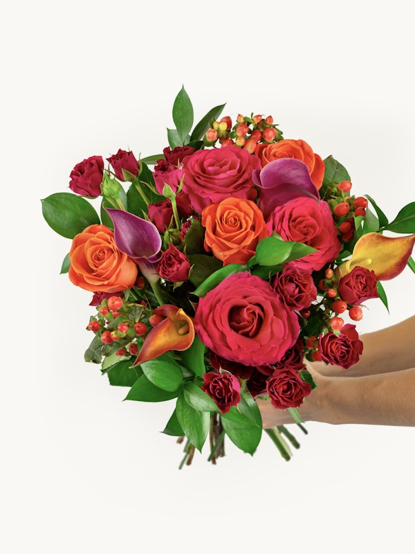 A lush bouquet with a mix of vibrant red roses, orange roses, and purple calla lilies, accented with green foliage, presented against a white background.