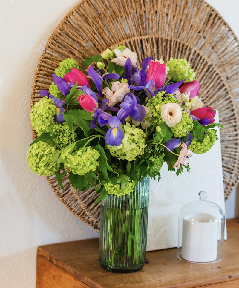 Vibrant bouquet of fresh flowers with purple tulips and blue irises, complemented by green hydrangeas, in a clear glass vase, set against a circular wicker background.