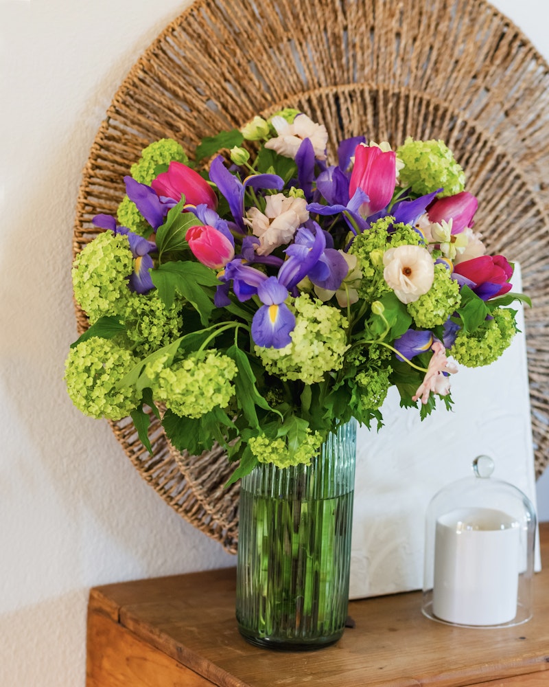 Vibrant bouquet of fresh flowers with purple tulips and blue irises, complemented by green hydrangeas, in a clear glass vase, set against a circular wicker background.
