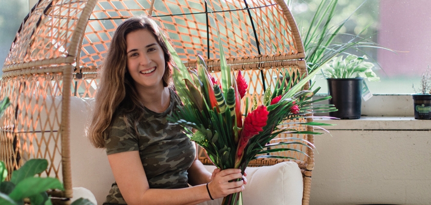 A smiling woman in a camo shirt holds a vibrant floral bouquet indoors, surrounded by greenery.