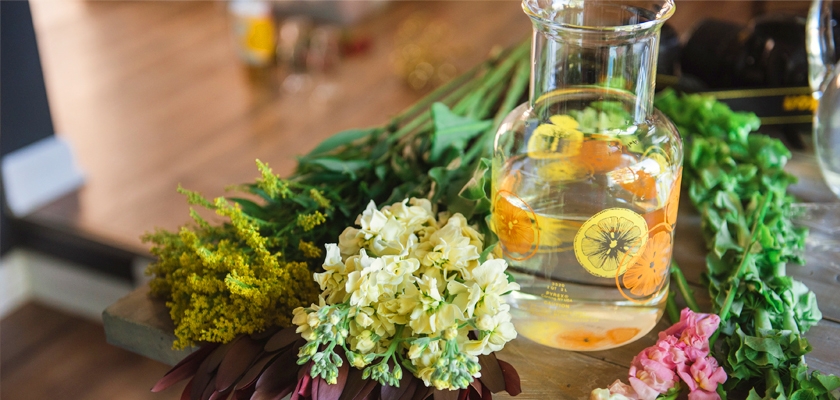 A colorful assortment of fresh flowers and herbs beside a clear vase with water and citrus slices.