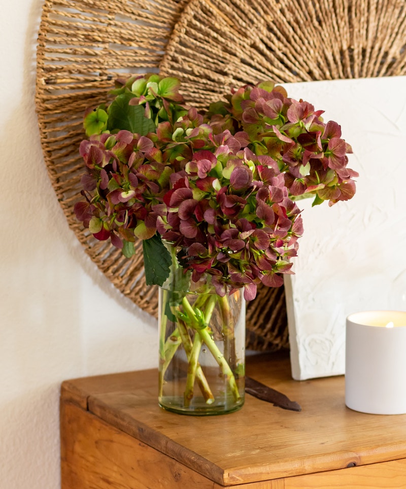 A bouquet of dried purple hydrangeas arranged in a clear glass vase on a wooden table, with decorative woven wall art and a lit candle in the background.