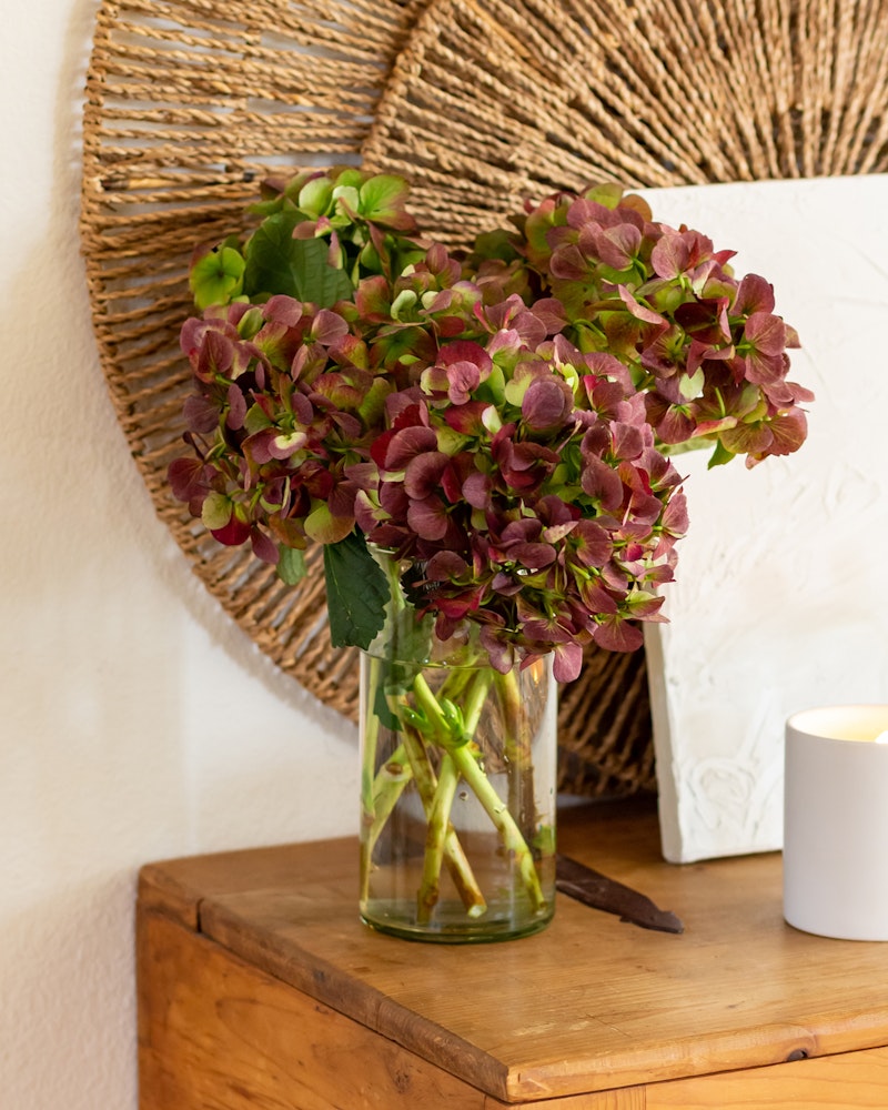 A bouquet of dried purple hydrangeas arranged in a clear glass vase on a wooden table, with decorative woven wall art and a lit candle in the background.