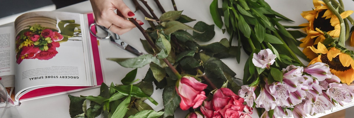 A person arranging a colorful bouquet of roses and sunflowers, with a floral guidebook nearby.