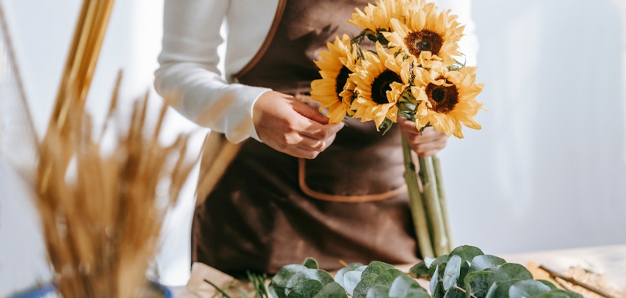 A florist holding a vibrant bouquet of sunflowers, surrounded by greenery and floral supplies.