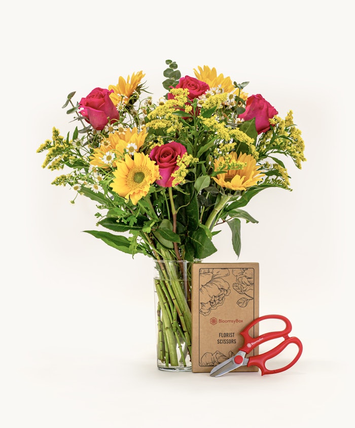 A vibrant bouquet featuring pink roses, sunflowers, and lush greenery in a stylish glass vase.