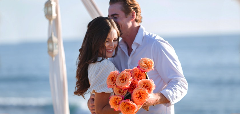 A joyful couple embracing on the beach, surrounded by vibrant orange roses, symbolizing love.