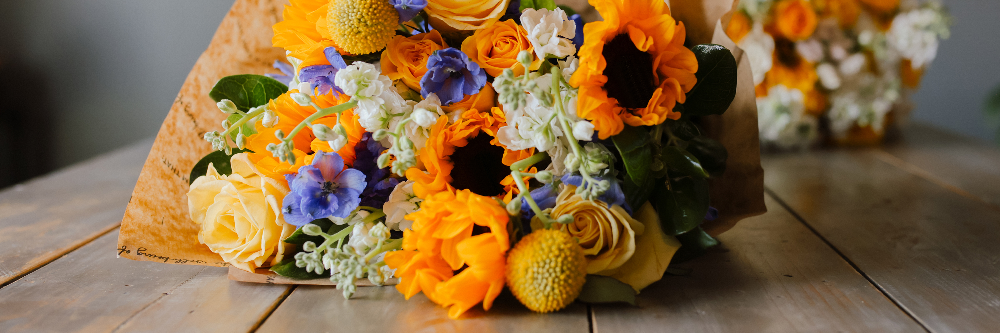 Vibrant floral bouquet featuring sunflowers, orange roses, and blue accents on a rustic table.