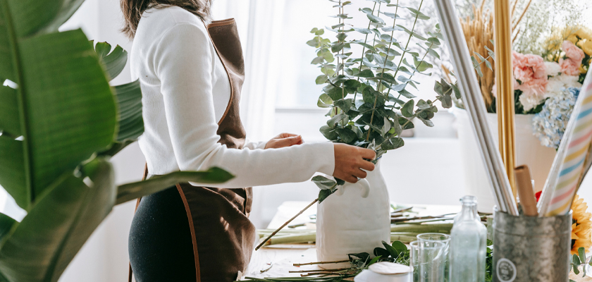 A florist arranging eucalyptus and green foliage in a minimalistic vase at a cozy workspace.