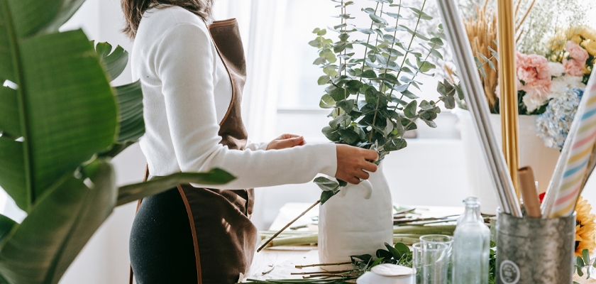 A florist arranging eucalyptus and green foliage in a minimalistic vase at a cozy workspace.