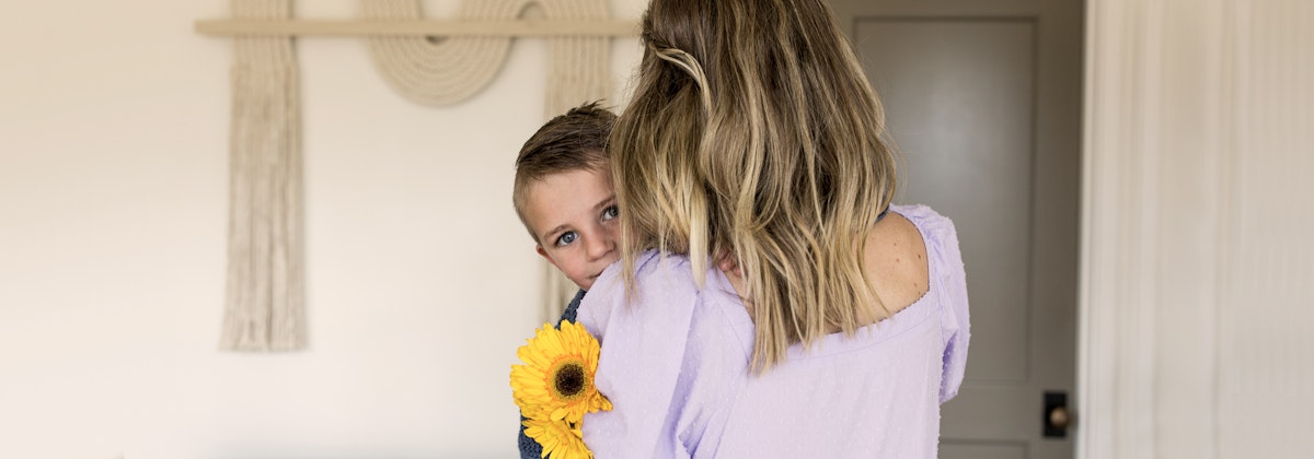 A mother holds her son who is peeking over her shoulder, surrounded by warm home decor.