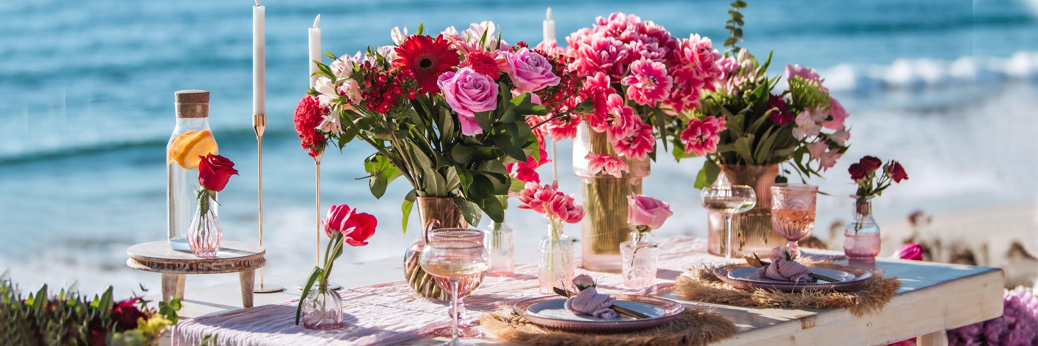 Elegant beachside table set with vibrant floral arrangements, candles, and charming decor.